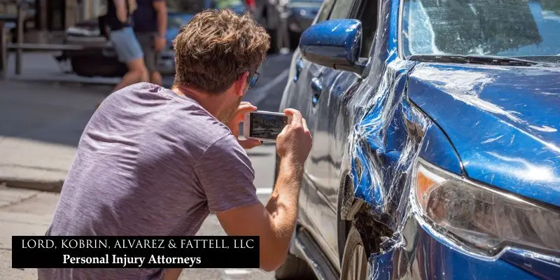 Driver photographs visible damage to rental car on NJ street after minor traffic accident for insurance purposes.